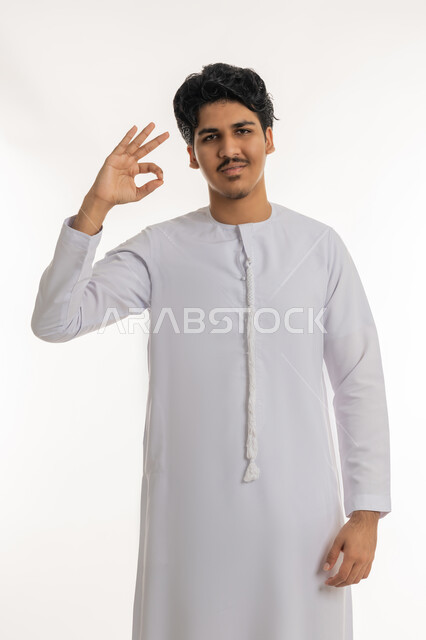 مفهوم الموافقة و القبول، علامة الامتياز و الجودة, النظر إلى الكاميرا بإيماءات السعادة و السرور، بورتريه لشاب مراهق عربي خليجي إماراتي مبتسم يرتدي الكندورة يرفع يده بتعابير الإعجاب و الرضى، خلفية بيضاء