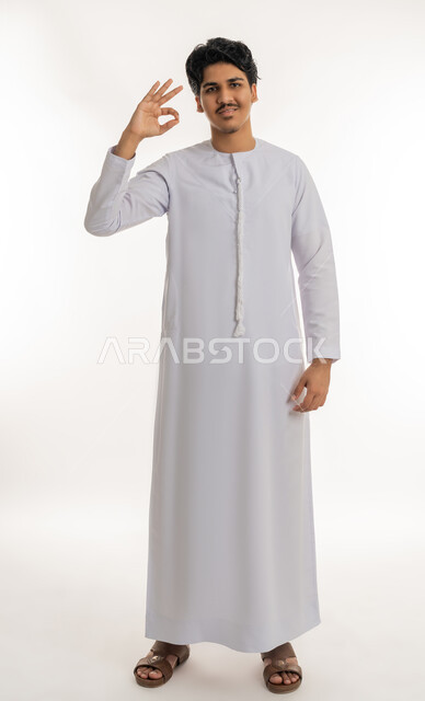 مفهوم الموافقة و القبول، علامة الامتياز و الجودة, النظر إلى الكاميرا بإيماءات السعادة و السرور، بورتريه لشاب مراهق عربي خليجي إماراتي مبتسم يرتدي الكندورة يرفع يده بتعابير الإعجاب و الرضى, الجسم بالطول الكامل، خلفية بيضاء