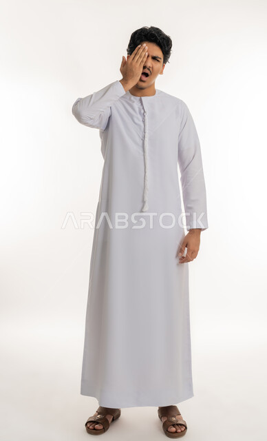 الالتهابات و المشاكل العينية و الحالات المرضية، الحاجة للفحص والعلاج، دخول شيء مزعج و مؤلم في العين, بورتريه لشاب مراهق عربي خليجي إماراتي يرتدي الكندورة يضع يده على عينه بتعابير الانزعاج و الالم, الجسم بالطول الكامل, خلفية بيضاء