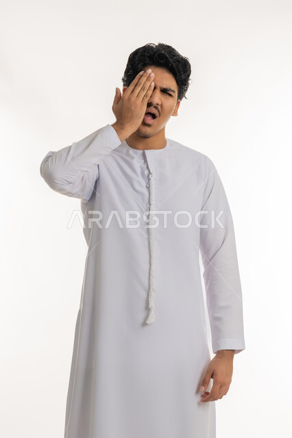 الالتهابات و المشاكل العينية و الحالات المرضية، الحاجة للفحص والعلاج، دخول شيء مزعج و مؤلم في العين, بورتريه لشاب مراهق عربي خليجي إماراتي يرتدي الكندورة يضع يده على عينه بتعابير الانزعاج و الالم, خلفية بيضاء