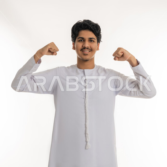 تحقيق الأهداف و إنجاز المهام, النظر بإيماءات السعادة و الفرح, بورتريه لشاب مراهق عربي خليجي إماراتي مبتسم يرتدي الكندورة يرفع يديه بتعابير القوة و النصر و الكسب, خلفية بيضاء