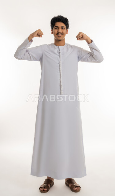تحقيق الأهداف و إنجاز المهام, النظر بإيماءات السعادة و الفرح, بورتريه لشاب مراهق عربي خليجي إماراتي مبتسم يرتدي الكندورة يرفع يديه بتعابير القوة و النصر و الكسب, الجسم بالطول الكامل, خلفية بيضاء