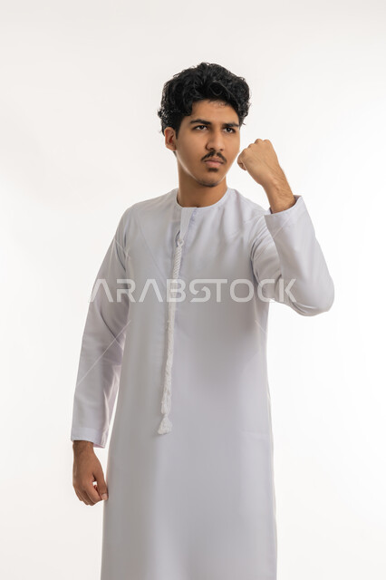 النظر بإيماءات الثقة بالنفس و الجدية و الحزم, مفهوم الاهتمام بالمظهر الخارجي, بورتريه لشاب مراهق عربي خليجي إماراتي يرتدي الكندورة يرفع يده بتعابير النصر و الفوز و القوة, خلفية بيضاء