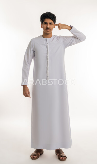 الإصرارا على الرأي و فعل ما يرغب, مفهوم التفكير والتحليل و الذكاء والفطنة, بورتريه لشاب مراهق عربي خليجي إماراتي يرتدي الكندورة يقف باستقامة و يشير بإصبع السبابة إلى رأسه بتعابير العناد, الجسم بالطول الكامل, خلفية بيضاء