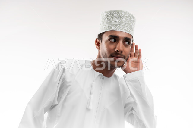 محاولة التجسس والتنصت, بورتريه مقرب لشاب عربي خليجي عماني يرتدي الدشداشة و الكمه يضع يده خلف اذنه بإيماءات تدل على الإنصات, حسن الإستماع و التركيز على الكلام, سماع الأسرار والمحادثات، خلفية بيضاء