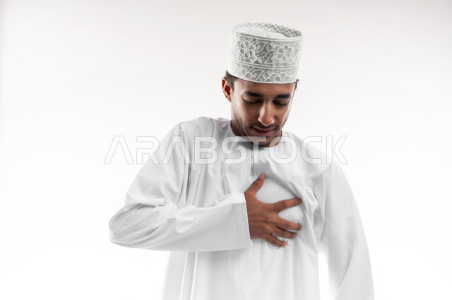 اضطرابات وأمراض قلبية, الاحساس بذبحه صدريه، بورتريه مقرب لشاب عربي خليجي عماني يرتدي الدشداشة و الكمه يضع يده على قلبه ويشعر بحالة سيئة من الوخز والألم, التعب والإرهاق والضغوطات النفسية, خلفية بيضاء