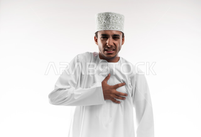 اضطرابات وأمراض قلبية, الاحساس بذبحه صدريه، بورتريه مقرب لشاب عربي خليجي عماني يرتدي الدشداشة و الكمه ينظر الى الكاميرا اليضع يده على قلبه ويشعر بحالة سيئة من الوخز والألم, التعب والإرهاق والضغوطات النفسية, خلفية بيضاء