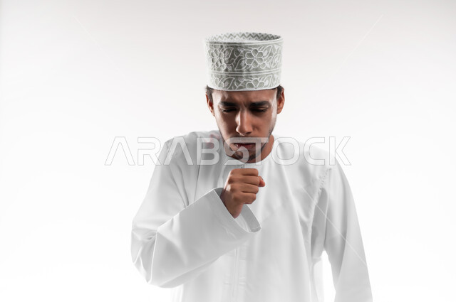 الشعور بالمرض والكحة، الانفلونزا والتهاب الرئة، الحاجة الى العلاج والدواء, بورتريه مقرب لشاب عربي خليجي عماني يرتدي الدشداشة و الكمه يضع يديه على فمه بإيماءات السعال, اعراض التعب والوهن، خلفية بيضاء