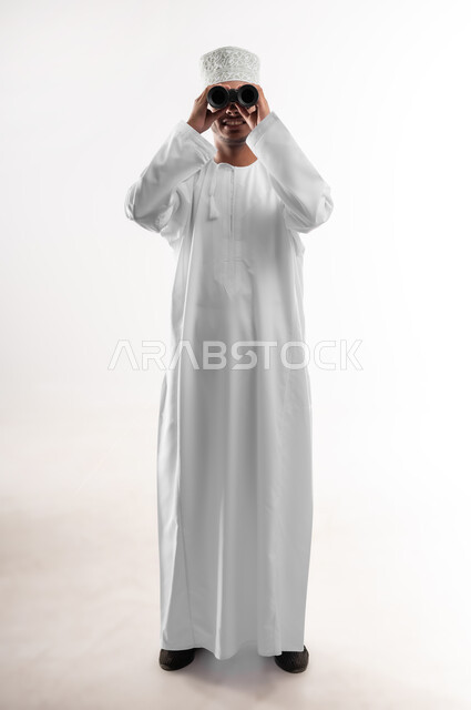 Seeing distant objects through binoculars, love of adventure and exploration, portrait of a young Arab Gulf Omani man wearing a dishdasha and a cloak looking through a binocular view (telescope) with gestures of joy and pleasure, future planning for goals and ambitions, full-length body image, white background