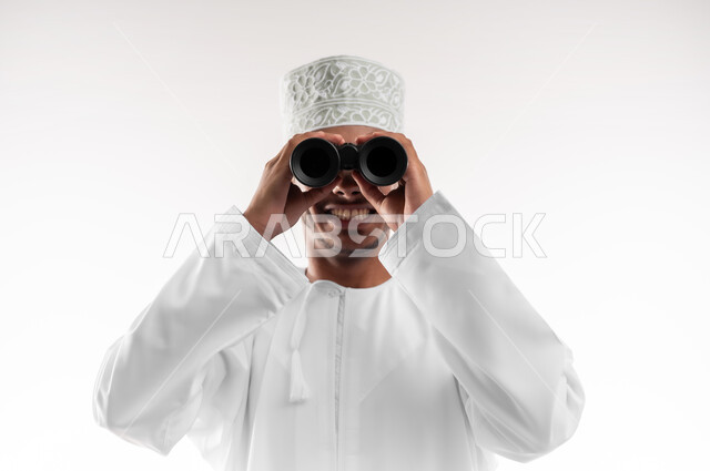 Future planning for goals and ambitions, seeing distant things through binoculars, love of adventure and exploration, close-up portrait of a young Gulf Arab Omani man wearing a dishdasha and a cloak looking through a binocular view (telescope) with gestures of joy and pleasure, white background