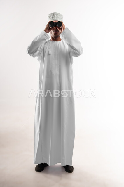 رؤية الاشياء البعيدة عبر المنظار، حب المغامرة والاستكشاف، بورتريه لشاب عربي خليجي عماني يرتدي الدشداشة والكمه ينظر من خلال المنظور ثنائي العينين (التلسكوب) بإيماءات الفرح والسرور، التخطيط المستقبلي للأهداف والطموحات، صورة الجسم بالطول الكامل، خلفية بيضاء