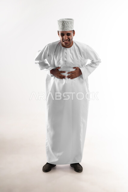 مفهوم أمراض والتهابات الجهاز الهضمي، الاشارة إلى المعدة بتعابير الإرهاق والتعب, بورتريه لشاب عربي خليجي عماني يرتدي الدشداشة والكمه يضع يده على بطنه بإيماءات الانزعاج والألم الشديد، صورة الجسم بالطول الكامل، خلفية بيضاء