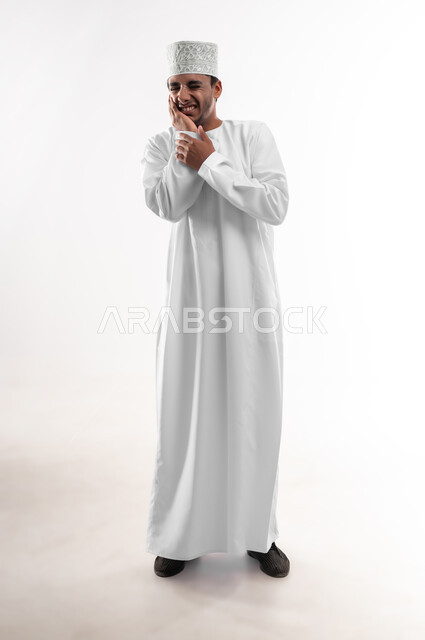 مشاكل التهاب اللثة وتسوس الأسنان, الاشارة إلى الفم بتعابير الإرهاق والتعب, بورتريه لشاب عربي خليجي عماني يرتدي الدشداشة والكمه يضع يده على فمه بإيماءات الانزعاج والألم الشديد، صورة الجسم بالطول الكامل، خلفية بيضاء