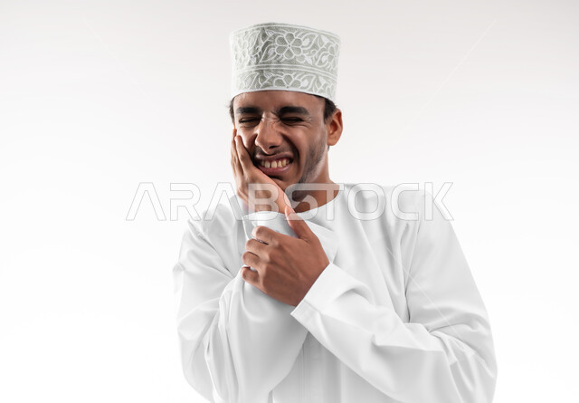 مشاكل التهاب اللثة وتسوس الأسنان, الاشارة إلى الفم بتعابير الإرهاق والتعب, بورتريه مقرب لشاب عربي خليجي عماني يرتدي الدشداشة والكمه يضع يده على فمه بإيماءات الانزعاج والألم الشديد، خلفية بيضاء