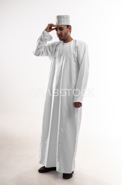 الإصابة بنوبات الشقيقة، إيماءات الصداع والإحساس بالمرض, الحاجه للعلاج وطلب المساعده, بورتريه لشاب عربي خليجي عماني يرتدي الدشداشة والكمه يمسك رأسه بيديه من شدة الألم, الشعور بالتعب والإرهاق, صورة الجسم بالطول الكامل، خلفية بيضاء