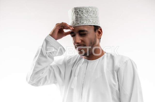 الإصابة بنوبات الشقيقة، إيماءات الصداع والإحساس بالمرض, الحاجه للعلاج وطلب المساعده, بورتريه مقرب لشاب عربي خليجي عماني يرتدي الدشداشة والكمه ينظر الى الكاميرا يمسك رأسه بيديه من شدة الألم, الشعور بالتعب والإرهاق, خلفية بيضاء