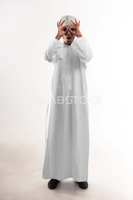 تعابير المشاهدة والتوضيح، مفهوم الرؤية المستقبلية، بورتريه لشاب عربي خليجي عماني يرتدي الدشداشة والكمه ينظر الى الكاميرا يضع يديه على عينيه بشكل عدسة بإيماءات الدهشة والذهول، الشعور بالفرح والسعادة، صورة الجسم بالطول الكامل، خلفية بيضاء