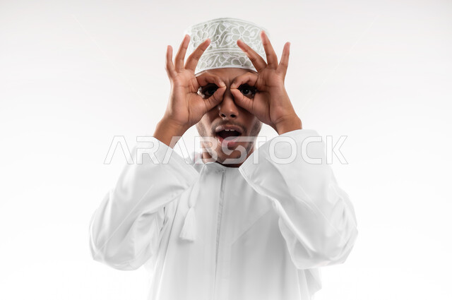 Expressions of observation and clarification, concept of future vision, close-up portrait of a young Gulf Arab Omani man wearing a dishdasha and a cloak looking at the camera, putting his hands over his eyes in the form of a lens with gestures of astonishment and amazement, feeling of joy and happiness, white background