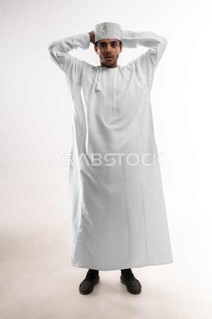 التفاجؤ والاستغراب من شيء ما، النظر إلى الكاميرا بإيماءات التعجب والدهشة، بورتريه لشاب عربي خليجي عماني يرتدي الدشداشة والكمه يضع يديه خلف رأسه يشعر بالصدمة والذهول والانبهار، تعابير النجاح وتحقيق الأهداف, صورة الجسم بالطول الكامل، خلفية بيضاء