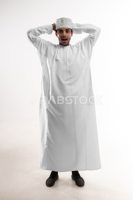 التفاجؤ والاستغراب من شيء ما، النظر إلى الكاميرا بإيماءات التعجب والدهشة، بورتريه لشاب عربي خليجي عماني يرتدي الدشداشة والكمه يضع يديه خلف رأسه يشعر بالصدمة والذهول والانبهار، تعابير النجاح وتحقيق الأهداف, صورة الجسم بالطول الكامل، خلفية بيضاء