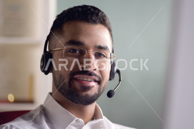 موظف عربي خليجي سعودي يعمل في خدمة العملاء ويرتدي سماعة الاتصال، استعلامات واستفسارات، الاجابه على اسئلة العملاء، اجراء اتصال فيديو من خلال الحاسوب، نظارة طبية، متابعة وتطوير العمل