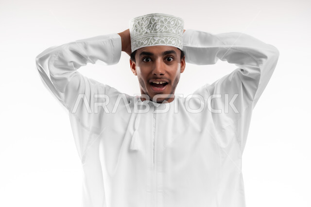 النظر إلى الكاميرا بإيماءات التعجب والدهشة، التفاجؤ والاستغراب من شيء ما، بورتريه مقرب لشاب عربي خليجي عماني يرتدي الدشداشة والكمه يضع يديه خلف رأسه يشعر بالصدمة والذهول والانبهار، تعابير النجاح وتحقيق الأهداف, خلفية بيضاء