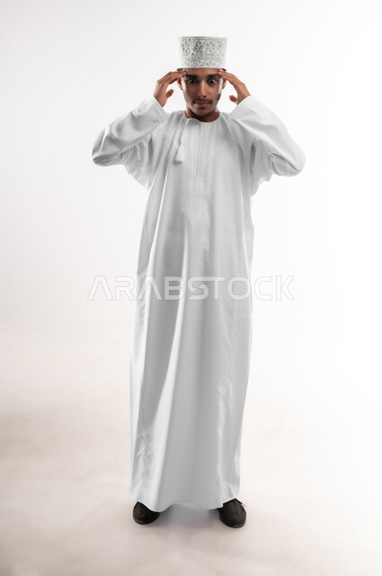 إيماءات الصداع والإحساس بالمرض, الحاجه للعلاج وطلب المساعده, بورتريه لشاب عربي خليجي عماني يرتدي الدشداشة والكمه ينظر الى الكاميرا يمسك رأسه بيديه من شدة الألم, الشعور بالتعب والإرهاق, الإصابة بنوبات الشقيقة، صورة الجسم بالطول الكامل، خلفية بيضاء