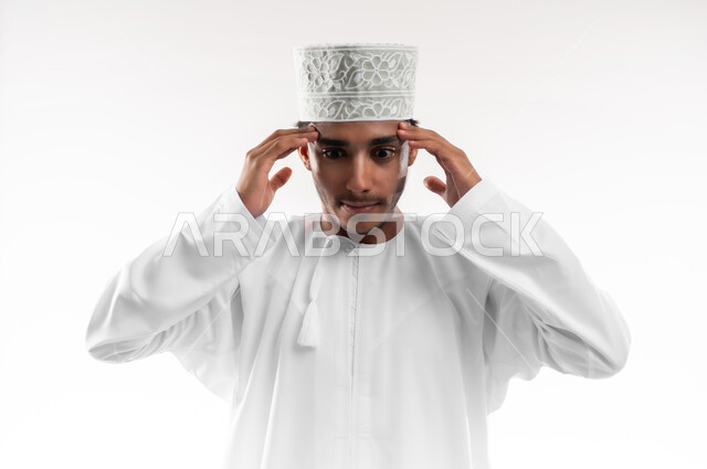 إيماءات الصداع والإحساس بالمرض, الحاجه للعلاج وطلب المساعده, بورتريه مقرب لشاب عربي خليجي عماني يرتدي الدشداشة والكمه ينظر الى الكاميرا يمسك رأسه بيديه من شدة الألم, الشعور بالتعب والإرهاق, الإصابة بنوبات الشقيقة، خلفية بيضاء