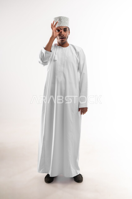 مفهوم الرؤية المستقبلية، تعابير المشاهدة والتوضيح، بورتريه لشاب عربي خليجي عماني يرتدي الدشداشة والكمه ينظر الى الكاميرا يضع يده على عينه بشكل عدسة بإيماءات الدهشة والذهول، الشعور بالفرح والسعادة، صورة الجسم بالطول الكامل، خلفية بيضاء