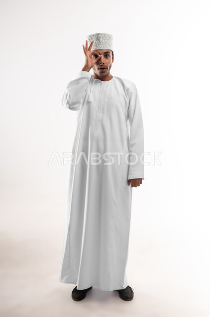 مفهوم الرؤية المستقبلية، تعابير المشاهدة والتوضيح، بورتريه لشاب عربي خليجي عماني يرتدي الدشداشة والكمه ينظر الى الكاميرا يضع يده على عينه بشكل عدسة بإيماءات الدهشة والذهول، الشعور بالفرح والسعادة، صورة الجسم بالطول الكامل، خلفية بيضاء