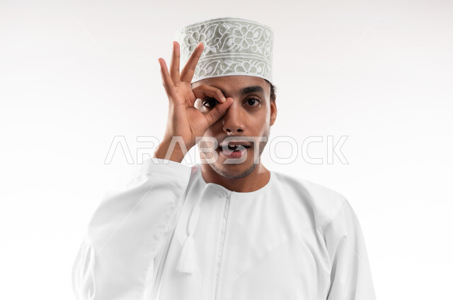 مفهوم الرؤية المستقبلية، تعابير المشاهدة والتوضيح، بورتريه مقرب لشاب عربي خليجي عماني يرتدي الدشداشة والكمه ينظر الى الكاميرا يضع يده على عينه بشكل عدسة بإيماءات الدهشة والذهول، الشعور بالفرح والسعادة، خلفية بيضاء