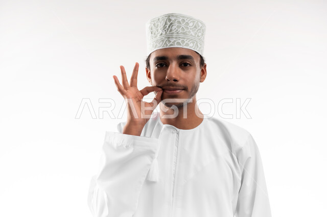 مفهوم الرجولة والقوة، التهديد والتوعد لشيء ما، بورتريه مقرب لشاب عربي خليجي عماني يرتدي الدشداشة والكمه ينظر الى الكاميرا يمسك شاربه بإيماءات الالتزام والوفاء بالوعود، خلفية بيضاء