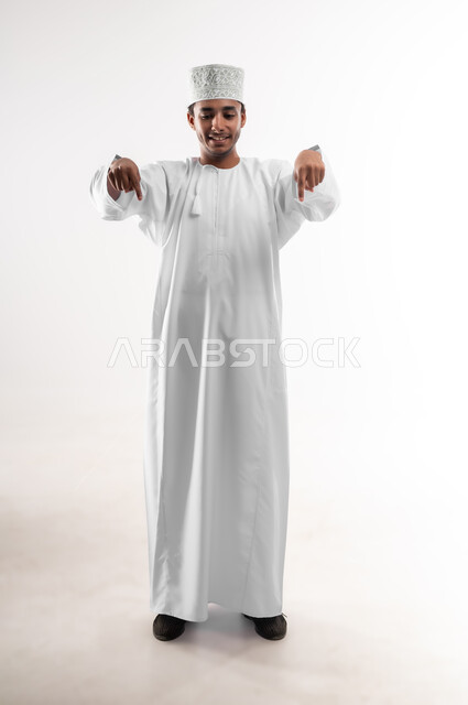 إشارات وحركات يدين تدل على هذا المكان (هنا)، تحديد موقع معين، بورتريه لشاب عربي خليجي عماني يرتدي الدشداشة والكمه يرفع يده للاعلى يشير بأصبع السبابة الى هدف محدد، مفهوم النقر والضغط والتفاعل مع العناصر الهولوجرامية، صورة الجسم بالطول الكامل، خلفية بيضاء