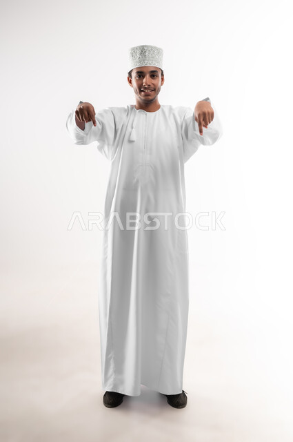 إشارات وحركات يدين تدل على هذا المكان (هنا)، تحديد موقع معين، بورتريه لشاب عربي خليجي عماني يرتدي الدشداشة والكمه يرفع يده للاعلى يشير بأصبع السبابة الى هدف محدد، مفهوم النقر والضغط والتفاعل مع العناصر الهولوجرامية، صورة الجسم بالطول الكامل، خلفية بيضاء