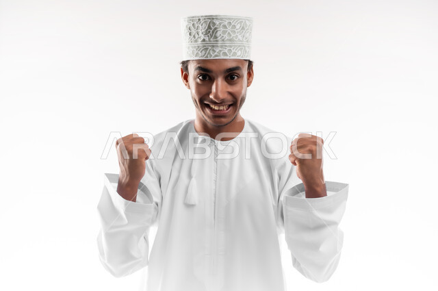 الانتصار والفوز والنجاح، مفهوم الانجاز وتحقيق الاهداف، بورتريه مقرب لشاب عربي خليجي عماني يرتدي الدشداشة والكمه ينظر الى الكاميرا يرفع يديه للاعلى بإيماءات الفرح والسرور, الشعور بالسعادة والقوة, خلفية بيضاء