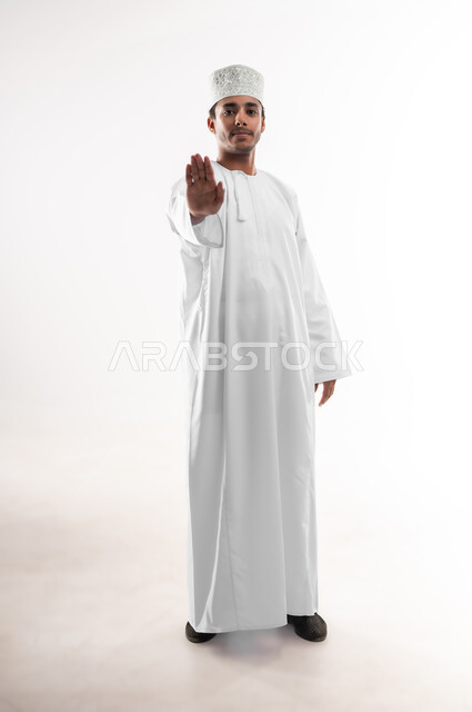 مفهوم الرفض والامتناع والانزعاج, ايماءات الحزم والجدية, طلب التوقف وعدم الاقتراب, بورتريه لشاب عربي خليجي عماني يرتدي الدشداشة والكمه ينظر الى الكاميرا يمد يده نحو الأمام مع رفع الكف بتعابير النهي عن فعل امر ما, صورة الجسم بالطول الكامل, خلفية بيضاء