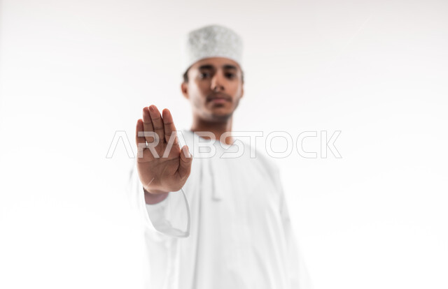 مفهوم الرفض والامتناع والانزعاج, ايماءات الحزم والجدية, طلب التوقف وعدم الاقتراب, بورتريه مقرب لشاب عربي خليجي عماني يرتدي الدشداشة والكمه ينظر الى الكاميرا يمد يده نحو الأمام مع رفع الكف بتعابير النهي عن فعل امر ما, خلفية بيضاء