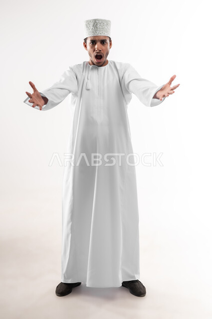 مفهوم الغضب والانفعال, بورتريه لشاب عربي خليجي عماني يرتدي الدشداشة و الكمه ينظر الى الكاميرا يرفع يديه عاليا بإيماءات التهيج و التوتر, تعابير الصراخ والضغط النفسي, صورة الجسم بالطول الكامل, خلفية بيضاء