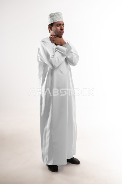 الشعور بالمرض والكحة، الانفلونزا والتهاب الرئة، الحاجة الى العلاج والدواء, بورتريه لشاب عربي خليجي عماني يرتدي الدشداشة و الكمه يضع يديه على رقبته بإيماءات ضيق التنفس, الشعور بالاختناق والتعب الشديد، صورة الجسم بالطول الكامل, خلفية بيضاء