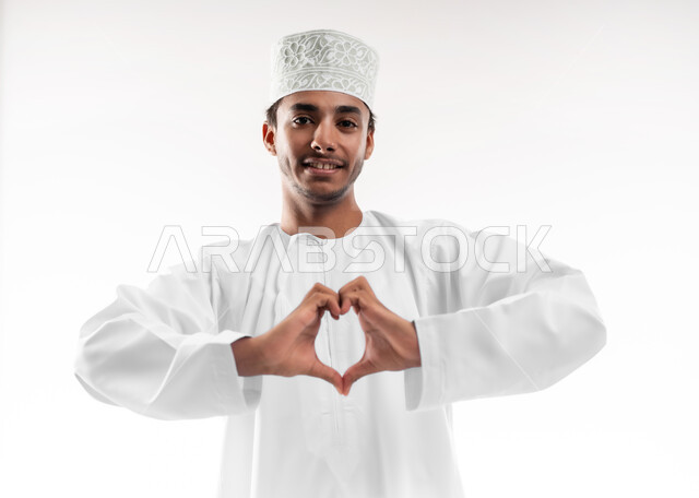 ايماءات يدين على شكل قلب، اظهار مشاعر الحب والشغف، مفهوم المحبة والمودة، بورتريه مقرب لشاب عربي خليجي عماني يرتدي الدشداشة والكمه ينظر إلى الكاميرا بتعابير السرور والسعادة، خلفية بيضاء