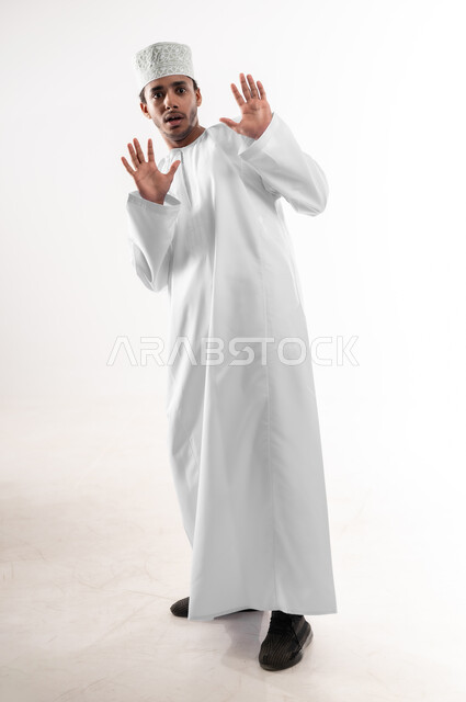 طلب التوقف والإمتناع عن فعل شيء ما، حماية النفس من ضرر خارجي, الرعب والذعر والرهبة، بورتريه لشاب عربي خليجي عماني يرتدي الدشداشة والكمه ينظر الى الكاميرا يرفع يديه للأعلى بإيماءات الخوف والصدمة، صورة الجسم بالطول الكامل، خلفية بيضاء