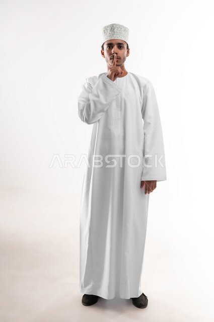 الهدوء والسكون وعدم الإزعاج، التوقف عن الكلام وخفض الصوت، بورتريه لشاب عربي خليجي عماني يرتدي الدشداشة والكمه ينظر الى الكاميرا يضع أصبع السبابة على فمه بإيماءات طلب السكوت والصمت، صورة الجسم بالطول الكامل, خلفية بيضاء