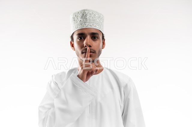 الهدوء والسكون وعدم الإزعاج، التوقف عن الكلام وخفض الصوت، بورتريه مقرب لشاب عربي خليجي عماني يرتدي الدشداشة والكمه ينظر الى الكاميرا يضع أصبع السبابة على فمه بإيماءات طلب السكوت والصمت، خلفية بيضاء