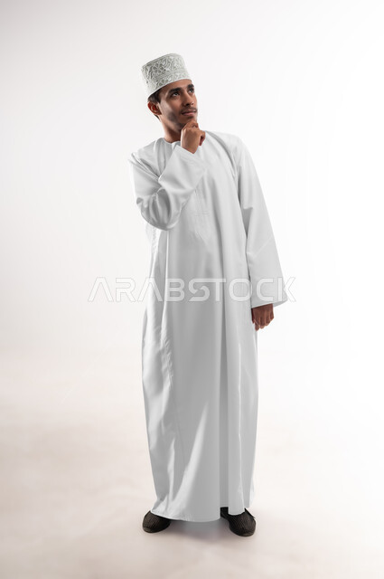 Contemplation, focus and planning for the future, finding a solution to a problem, portrait of a young Arab Gulf Omani man wearing a dishdasha and a sleeve, putting his hand under his chin with gestures of contemplation and thinking, the concept of absent-mindedness, full-length body image, white background