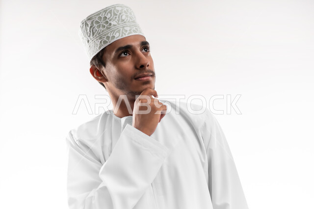 Contemplation, focus and planning for the future, finding a solution to a problem, close-up portrait of a young Arab Gulf Omani man wearing a dishdasha and a turban, putting his hand under his chin with gestures of contemplation and thinking, the concept of absent-mindedness, white background