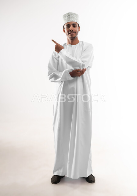 Positioning, portrait of a smiling young Omani Arab man wearing a dishdasha and a sleeve looking at the camera raising his hand up pointing with his index finger at a specific target, concept of clicking, pressing and interacting with holographic elements, full-length body image, white background