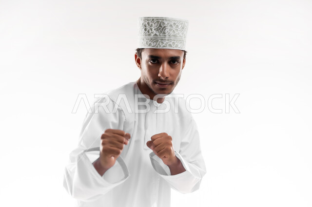 ممارسة رياضة الدفاع عن النفس، زيادة قدرة العضلات على التحمل، إتقان فنون الملاكمة، بورتريه مقرب لشاب عربي خليجي عماني يرتدي الدشداشة و الكمه ينظر الى الكاميرا يرفع قبضات يديه بإيماءات القوة، خلفية بيضاء