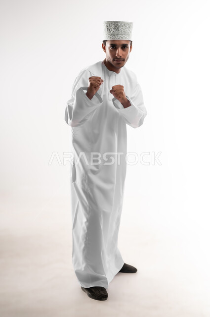 ممارسة رياضة الدفاع عن النفس، زيادة قدرة العضلات على التحمل، إتقان فنون الملاكمة، بورتريه لشاب عربي خليجي عماني يرتدي الدشداشة و الكمه ينظر الى الكاميرا يرفع قبضات يديه بإيماءات القوة، صورة الجسم بالطول الكامل، خلفية بيضاء