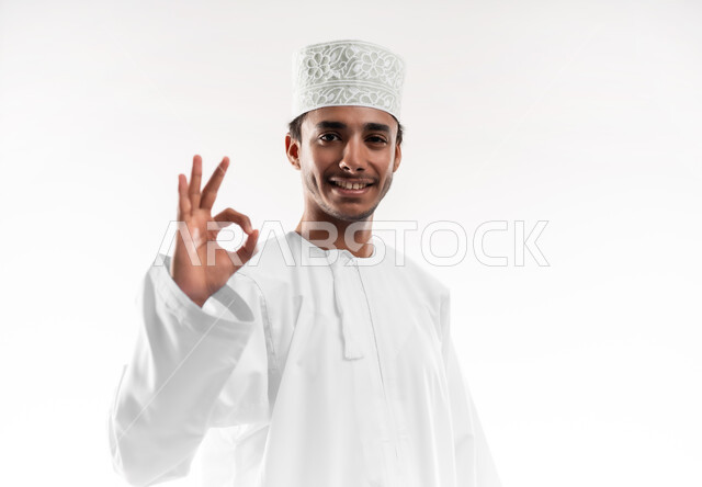 مفهوم القبول والموافقه، علامة الجوده و الإمتياز والتميز, بورتريه مقرب لشاب عربي خليجي عماني مبتسم يرتدي الدشداشة والكمه ينظر الى الكاميرا يرفع يديه للاعلى بإيماءات الإعجاب والرضا، خلفية بيضاء