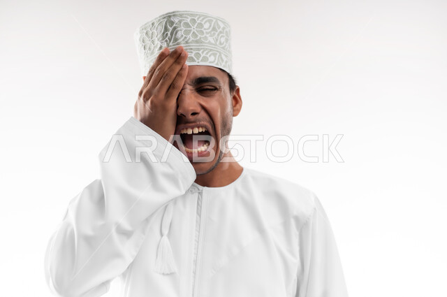 عدم القدرة على فتح العينين، مشاكل عينيه وحالات مرضية، الحاجة للفحص والعلاج، التهاب القرنية العينية، بورتريه مقرب لشاب عربي خليجي عماني يرتدي الدشداشة والكمه يضع يده على عينه بإيماءات الألم, خلفية بيضاء